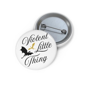 May include: White button pin with the text "Violent Little Thing" in black script. A black dragon and a gold dragon are featured below the text. The pin is round and has a metal backing.