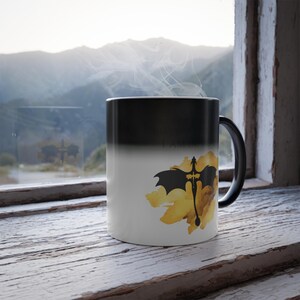 May include: A black and white color-changing mug with a dragon and sword design. The mug transitions from black to white when hot, revealing the image. The text "I AM FIRE" is also visible.