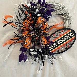 May include: A white twig wreath with a black and orange bow and a black and white sign that says "Happy Halloween". The wreath is decorated with white flowers, purple leaves, and black feathers.