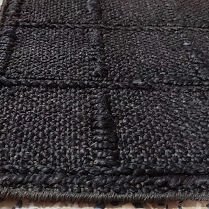 May include: A dark brown, rectangular rug featuring a grid pattern created by raised, woven fiber lines. The surface has a coarse, textured appearance with a thick, braided border edge that frames the repeating square sections across the rug.