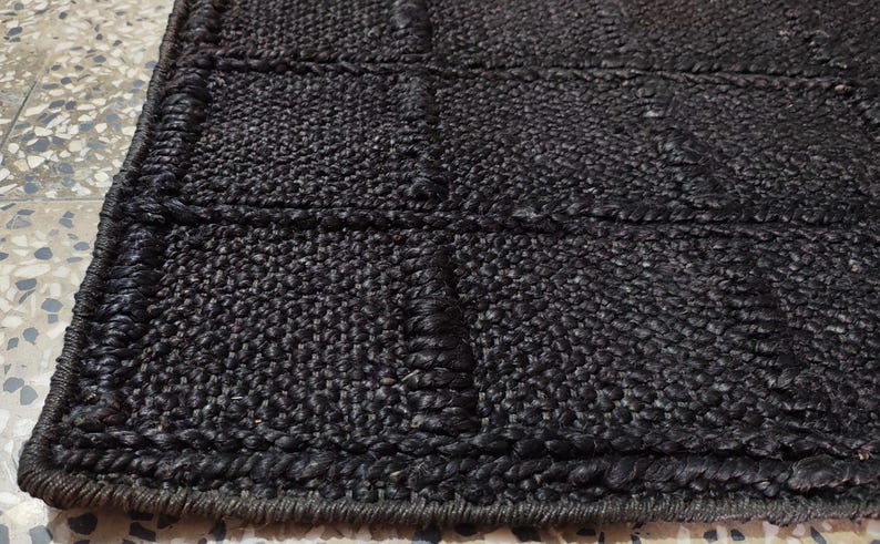 May include: A dark brown, rectangular rug featuring a grid pattern created by raised, woven fiber lines. The surface has a coarse, textured appearance with a thick, braided border edge that frames the repeating square sections across the rug.