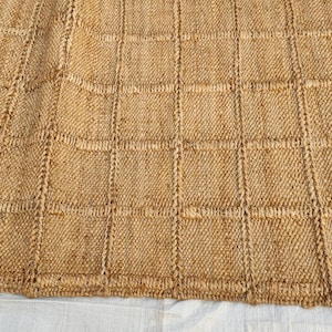 May include: A rectangular area rug crafted from natural tan fibers, featuring a prominent grid pattern created by raised, woven lines that form uniform squares across the entire surface of the textured, earthy-toned floor covering.