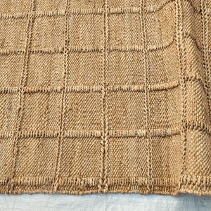 May include: A tan woven rug featuring a grid pattern of raised square sections. The texture is coarse and fibrous, with a natural light brown color throughout. The design consists of repeating squares separated by vertical and horizontal lines.