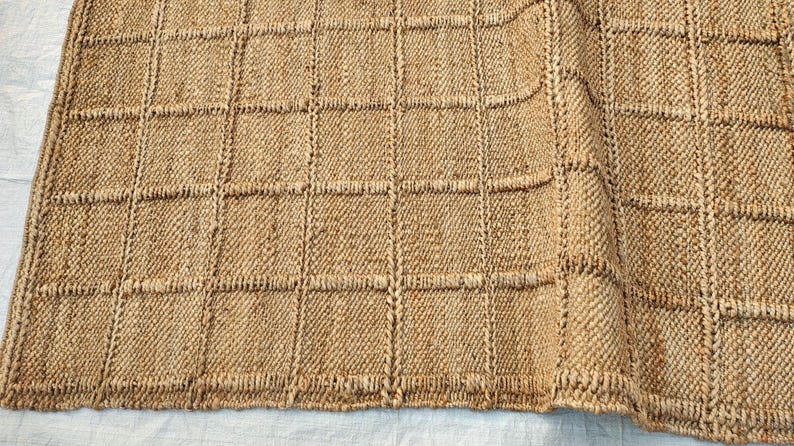 May include: A tan woven rug featuring a grid pattern of raised square sections. The texture is coarse and fibrous, with a natural light brown color throughout. The design consists of repeating squares separated by vertical and horizontal lines.