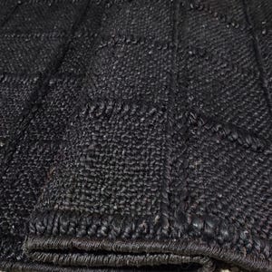 May include: A black area rug featuring a textured, woven surface with a grid pattern of raised lines. The rug is folded over at one corner, revealing the thickness and the consistent dark material used throughout the entire woven construction.