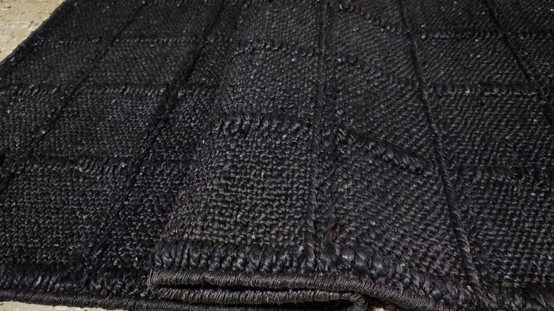May include: A black area rug featuring a textured, woven surface with a grid pattern of raised lines. The rug is folded over at one corner, revealing the thickness and the consistent dark material used throughout the entire woven construction.