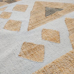 May include: A woven area rug featuring a light gray base with a repeating geometric pattern of tan diamonds and triangles. The texture is coarse and fibrous, with dark brown and tan striped borders visible along the edges of the fabric surface.