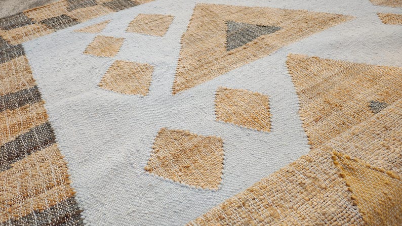 May include: A woven area rug featuring a light gray base with a repeating geometric pattern of tan diamonds and triangles. The texture is coarse and fibrous, with dark brown and tan striped borders visible along the edges of the fabric surface.