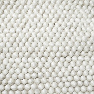 Puede incluir: Una alfombra blanca y esponjosa hecha de pequeñas bolas redondas de hilo. La alfombra tiene una superficie texturizada y un patrón sutil.