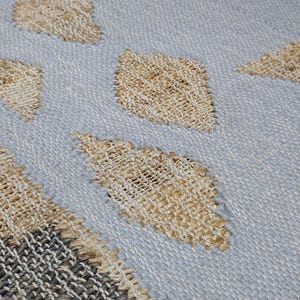 May include: A woven textile rug featuring a light blue base color with a repeating pattern of tan diamond shapes. The left side of the rug displays a border section with a grid-like weave pattern in alternating shades of dark gray and tan fibers.