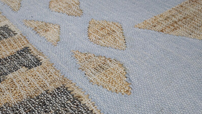 May include: A woven textile rug featuring a light blue base color with a repeating pattern of tan diamond shapes. The left side of the rug displays a border section with a grid-like weave pattern in alternating shades of dark gray and tan fibers.