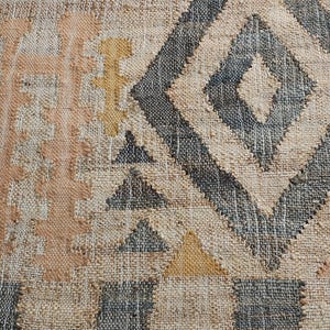 May include: A woven rug featuring a textured beige base with a geometric pattern in charcoal gray, muted peach, and mustard yellow. The design includes large diamonds, triangles, and vertical columns of stacked shapes across the fabric surface.
