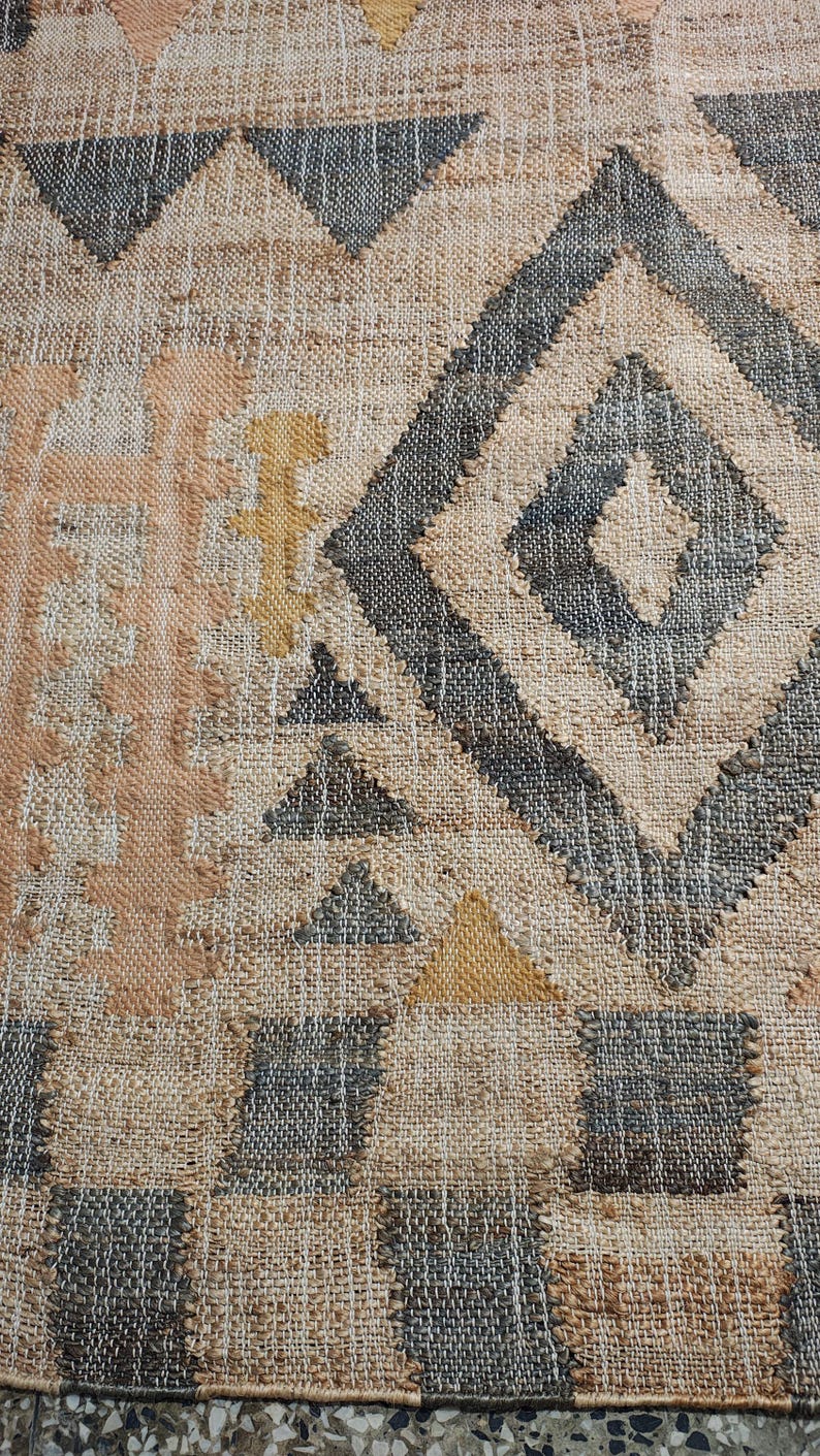 May include: A woven rug featuring a textured beige base with a geometric pattern in charcoal gray, muted peach, and mustard yellow. The design includes large diamonds, triangles, and vertical columns of stacked shapes across the fabric surface.