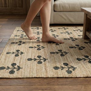 May include: A rectangular area rug made of natural tan woven fibers featuring a repeating dark gray floral pattern. The rug has a textured surface and is laid flat on a wooden floor, showcasing a consistent botanical print across the entire weave.