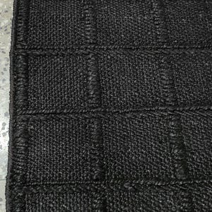 Puede incluir: Una alfombra negra tejida con una superficie texturizada y forma rectangular. La alfombra presenta un patr&oacute;n de rejilla con secciones rectangulares en relieve. El material parece ser una fibra natural, creando una est&eacute;tica r&uacute;stica. Para uso en interiores.