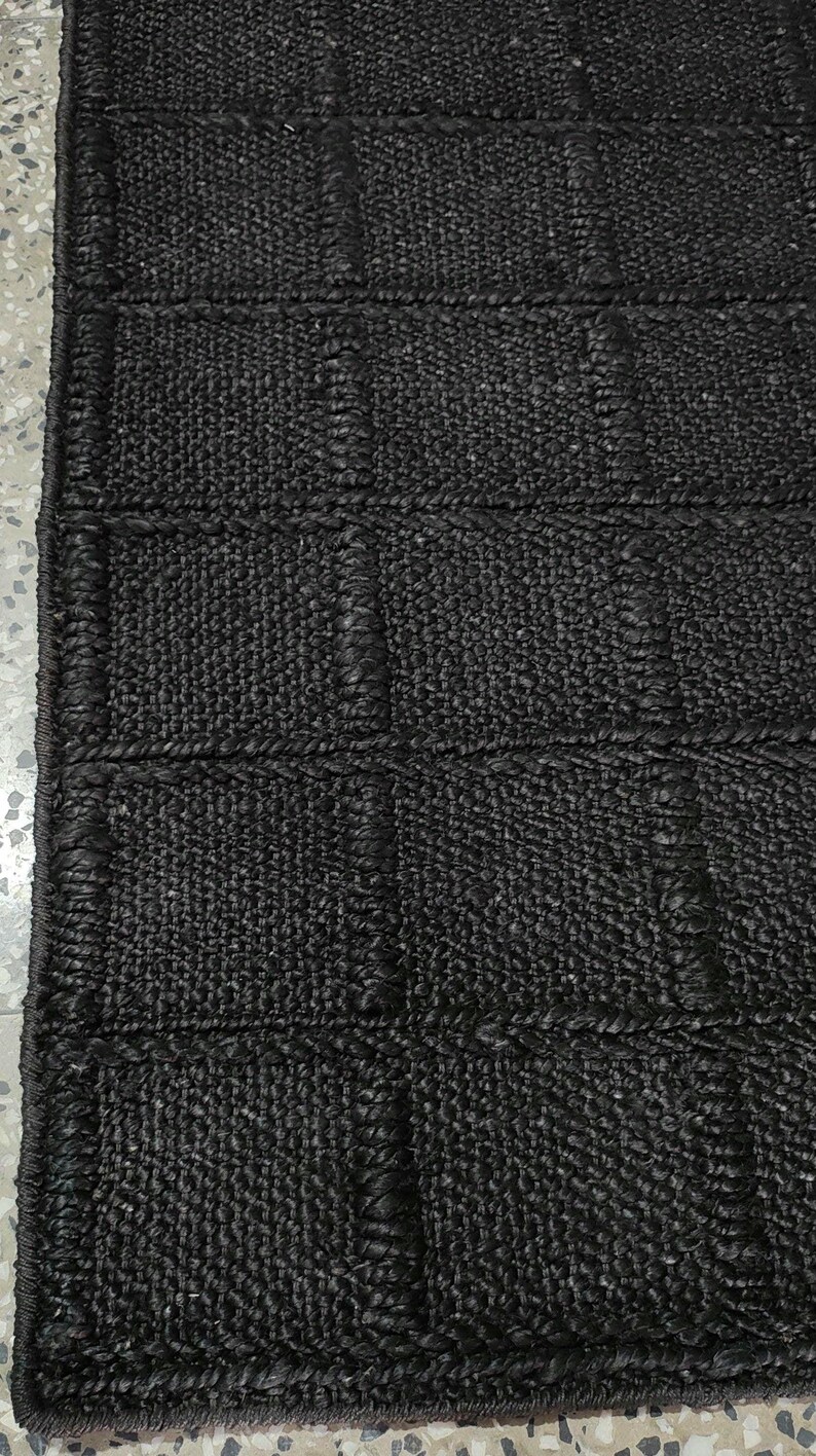 Puede incluir: Una alfombra negra tejida con una superficie texturizada y forma rectangular. La alfombra presenta un patr&oacute;n de rejilla con secciones rectangulares en relieve. El material parece ser una fibra natural, creando una est&eacute;tica r&uacute;stica. Para uso en interiores.