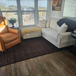 May include: A living room set featuring a tan leather recliner, a white sofa with a dark throw blanket, a white ottoman with a yellow knit throw, and a dark brown textured area rug. A small round side table and a black pedestal table are included.