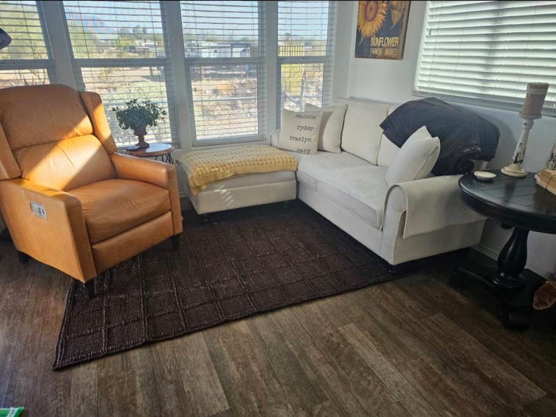 May include: A living room set featuring a tan leather recliner, a white sofa with a dark throw blanket, a white ottoman with a yellow knit throw, and a dark brown textured area rug. A small round side table and a black pedestal table are included.