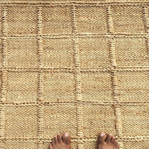 May include: A rectangular area rug crafted from natural tan jute fibers, featuring a prominent grid pattern created by raised, thicker woven lines that form uniform squares across the entire surface of the textured, earthy-toned floor covering.