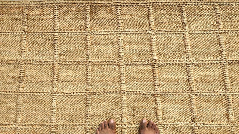 May include: A rectangular area rug crafted from natural tan jute fibers, featuring a prominent grid pattern created by raised, thicker woven lines that form uniform squares across the entire surface of the textured, earthy-toned floor covering.