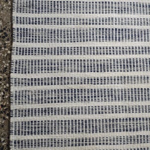 May include: A rectangular woven rug featuring a repeating pattern of thin, dark blue horizontal stripes set against a textured off-white background. The fabric displays a visible, dense weave structure with consistent, parallel lines throughout.