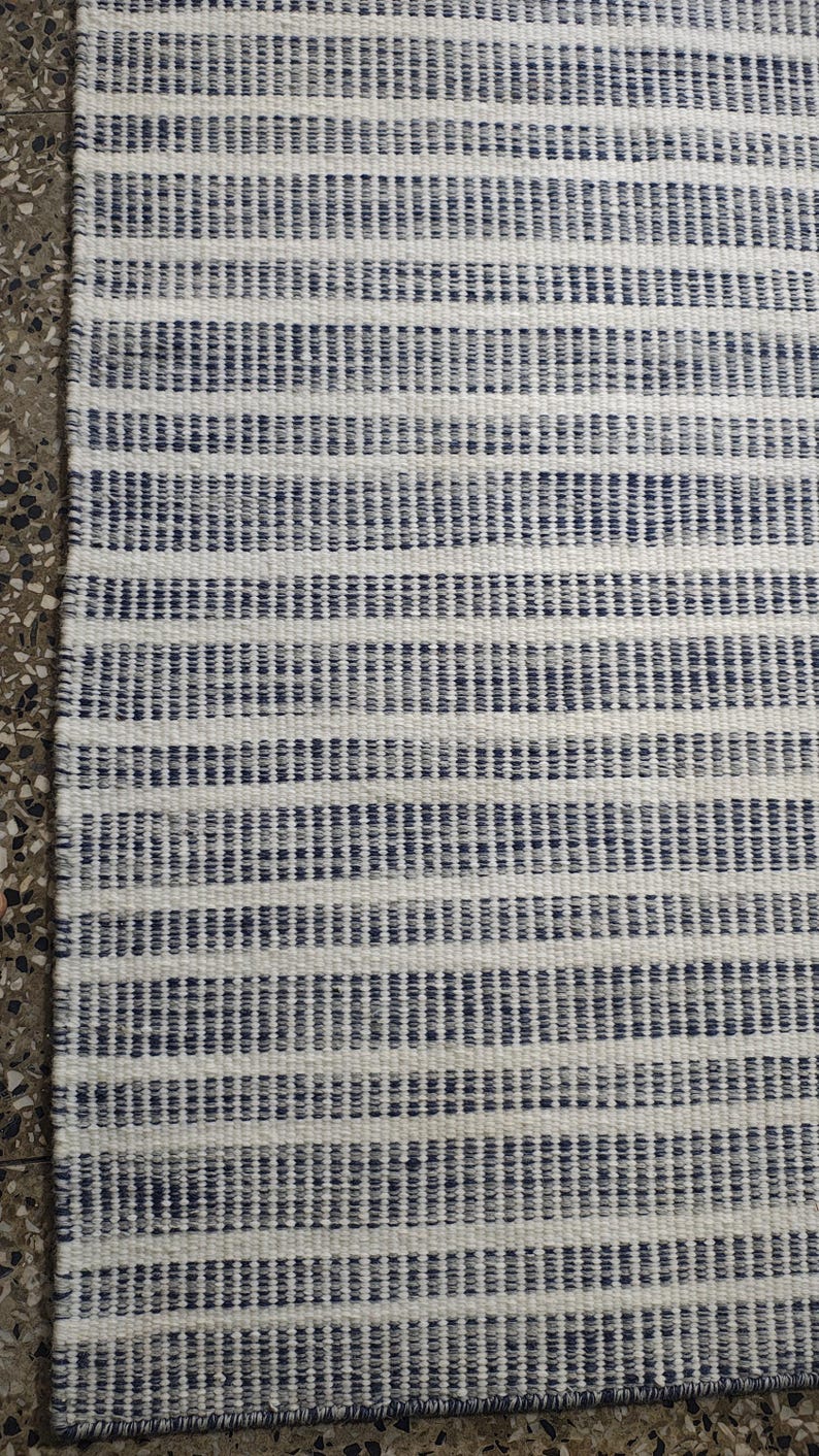 May include: A rectangular woven rug featuring a repeating pattern of thin, dark blue horizontal stripes set against a textured off-white background. The fabric displays a visible, dense weave structure with consistent, parallel lines throughout.