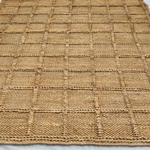 May include: A rectangular area rug crafted from natural tan fibers, featuring a prominent grid pattern created by raised, woven lines that form uniform squares across the entire surface of the textured, earthy-toned floor covering.