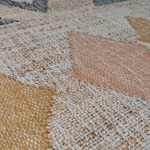 May include: A woven area rug featuring a repeating geometric diamond pattern in shades of mustard yellow, soft peach, and charcoal gray against a natural beige textured background. The fabric has a visible coarse weave with a tactile surface.