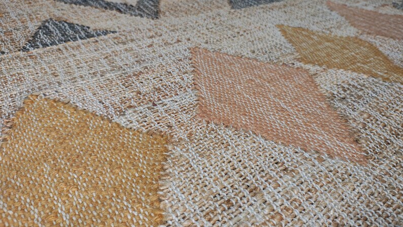 May include: A woven area rug featuring a repeating geometric diamond pattern in shades of mustard yellow, soft peach, and charcoal gray against a natural beige textured background. The fabric has a visible coarse weave with a tactile surface.