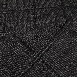Puede incluir: Primer plano de una alfombra negra tejida con una superficie texturizada. La alfombra presenta un patr&oacute;n similar a una cuadr&iacute;cula con secciones rectangulares elevadas. El color oscuro y la textura tejida sugieren un revestimiento de suelo duradero y elegante.