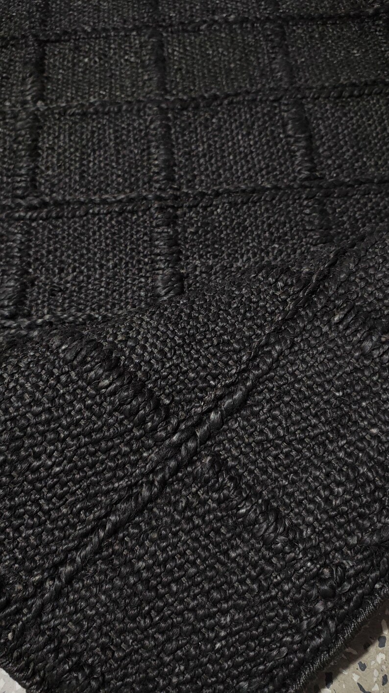 Puede incluir: Primer plano de una alfombra negra tejida con una superficie texturizada. La alfombra presenta un patr&oacute;n similar a una cuadr&iacute;cula con secciones rectangulares elevadas. El color oscuro y la textura tejida sugieren un revestimiento de suelo duradero y elegante.