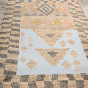 May include: A rectangular woven rug featuring a natural tan fiber base with a geometric pattern. The design includes a large light blue central block, dark gray and yellow diamond shapes, and a border of alternating dark gray and tan rectangular blocks.