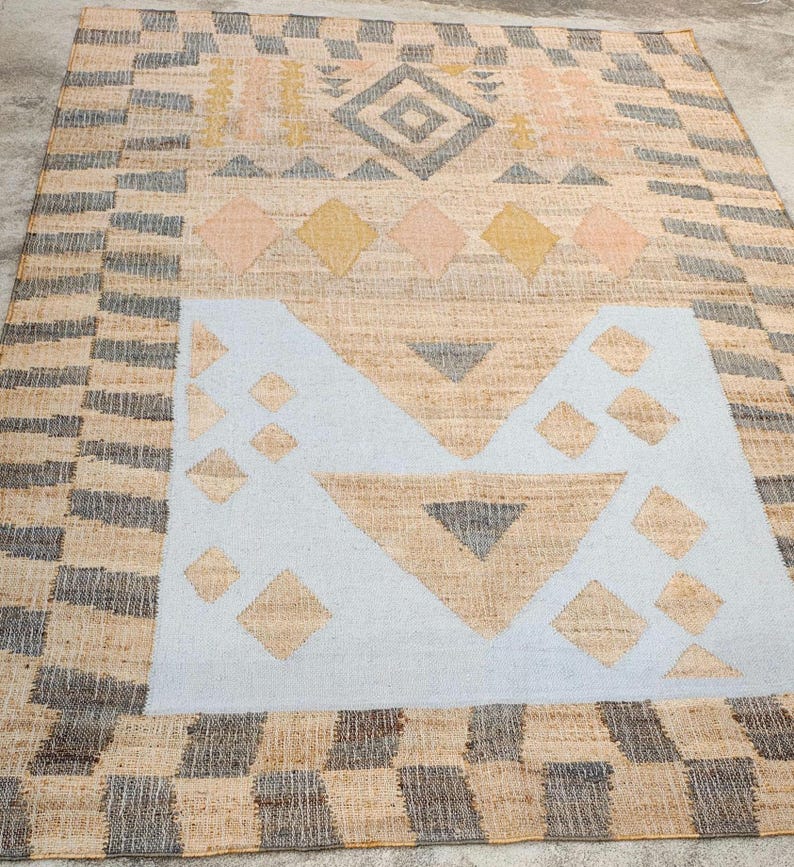 May include: A rectangular woven rug featuring a natural tan fiber base with a geometric pattern. The design includes a large light blue central block, dark gray and yellow diamond shapes, and a border of alternating dark gray and tan rectangular blocks.