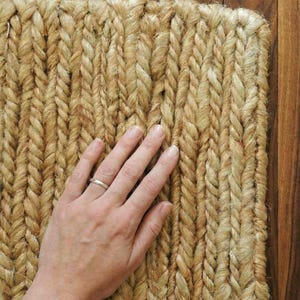 Puede incluir: Primer plano de una alfombra de yute tejida con una textura trenzada. La alfombra es de color beige natural, con variaciones de tono. La textura es visible y la alfombra parece hecha a mano. La alfombra está colocada sobre una superficie de madera.