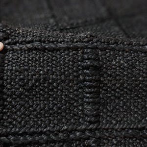 Puede incluir: Primer plano de una alfombra negra tejida con una superficie texturizada. La alfombra est&aacute; hecha de fibras gruesas y oscuras, y los bordes est&aacute;n cuidadosamente acabados. El patr&oacute;n de tejido crea un dise&ntilde;o similar a una cuadr&iacute;cula.