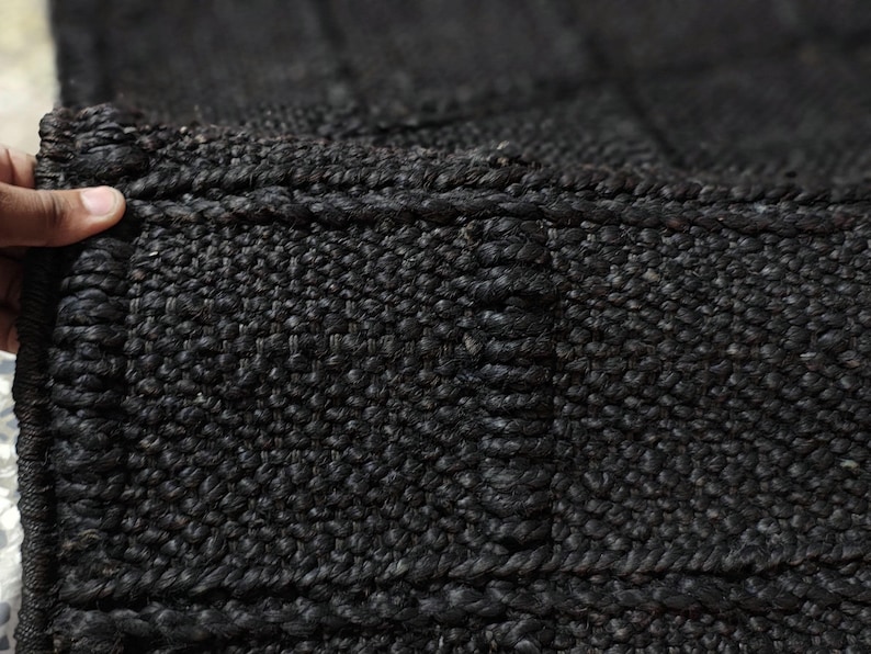 Puede incluir: Primer plano de una alfombra negra tejida con una superficie texturizada. La alfombra est&aacute; hecha de fibras gruesas y oscuras, y los bordes est&aacute;n cuidadosamente acabados. El patr&oacute;n de tejido crea un dise&ntilde;o similar a una cuadr&iacute;cula.