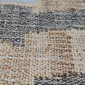 May include: A woven area rug featuring a checkered pattern composed of alternating squares in natural tan jute and dark charcoal gray fibers. The textile has a thick, textured weave with a visible grid structure and a finished bound edge.