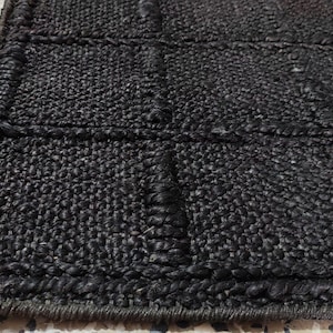 Puede incluir: Primer plano de una alfombra de yute negro tejida. La alfombra tiene un patr&oacute;n rectangular con bordes elevados y detalles de costura. El color oscuro y la superficie texturizada crean una est&eacute;tica r&uacute;stica y natural.