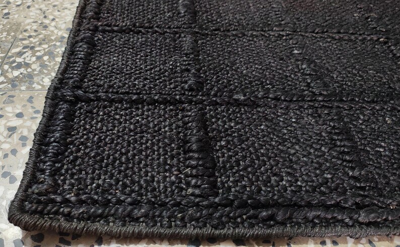 Puede incluir: Primer plano de una alfombra de yute negro tejida. La alfombra tiene un patr&oacute;n rectangular con bordes elevados y detalles de costura. El color oscuro y la superficie texturizada crean una est&eacute;tica r&uacute;stica y natural.