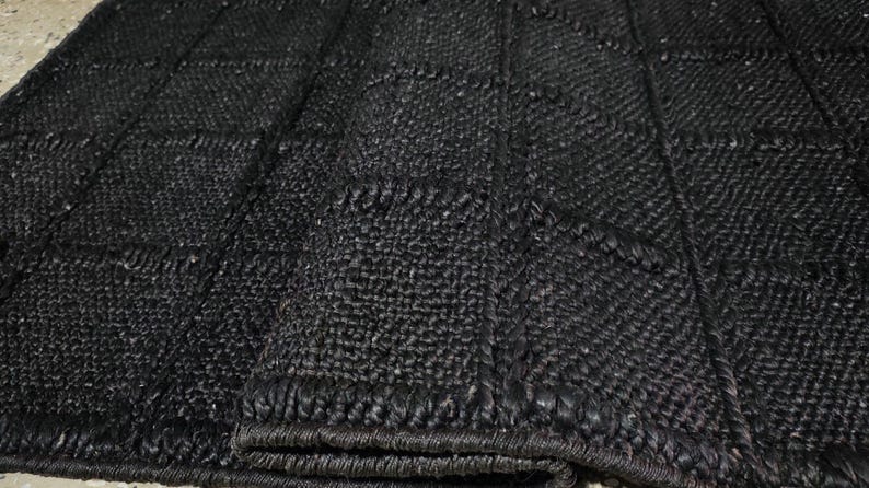 Puede incluir: Primer plano de una alfombra negra tejida con un patr&oacute;n geom&eacute;trico en forma de cuadr&iacute;cula. La alfombra tiene una superficie texturizada y un color oscuro, carb&oacute;n. Los bordes est&aacute;n cuidadosamente acabados con un borde tejido. Esta alfombra es adecuada para la decoraci&oacute;n del hogar.