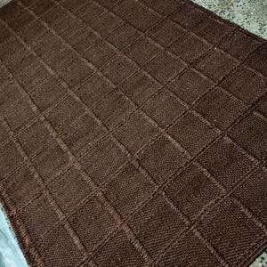 May include: A rectangular brown rug featuring a textured grid pattern composed of small, raised square sections. The rug has a consistent dark brown color throughout its surface, with visible knit-like stitching forming the square grid design.