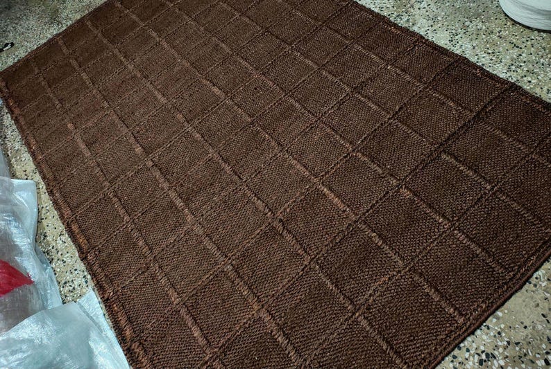 May include: A rectangular brown rug featuring a textured grid pattern composed of small, raised square sections. The rug has a consistent dark brown color throughout its surface, with visible knit-like stitching forming the square grid design.