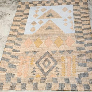 May include: A rectangular woven rug featuring a natural tan base with a border of alternating gray and tan blocks. The center displays a light blue panel with tan triangles and diamonds, alongside rows of yellow, pink, and gray geometric shapes.