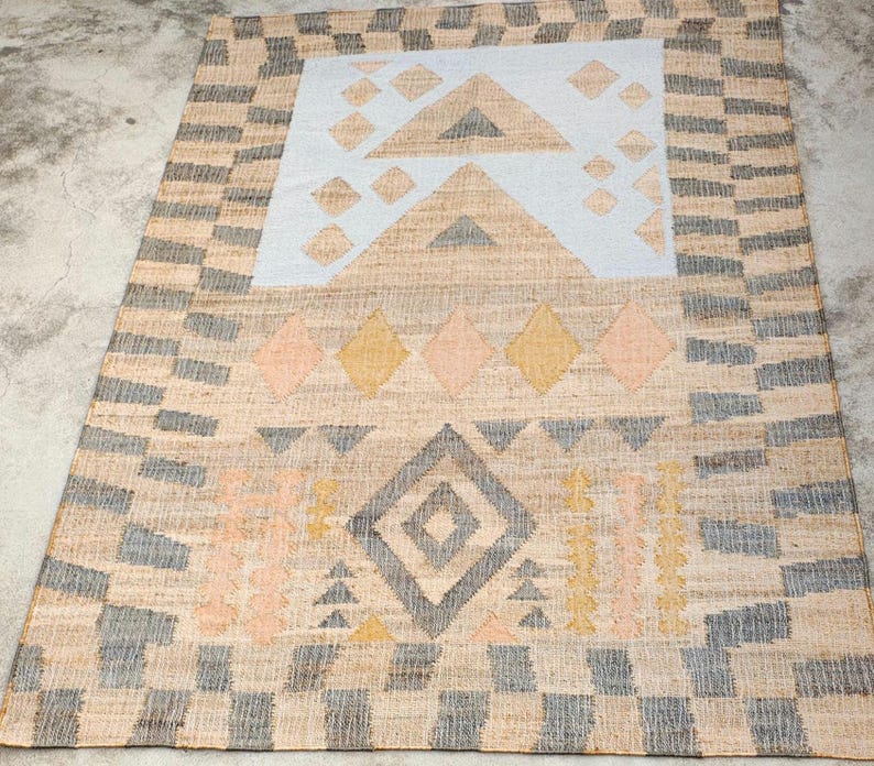 May include: A rectangular woven rug featuring a natural tan base with a border of alternating gray and tan blocks. The center displays a light blue panel with tan triangles and diamonds, alongside rows of yellow, pink, and gray geometric shapes.