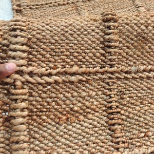 May include: A tan woven rug featuring a textured surface with a grid pattern created by thick, twisted fiber ropes. The material has a natural, earthy brown tone with a dense, knotted construction that forms a repeating square design throughout.