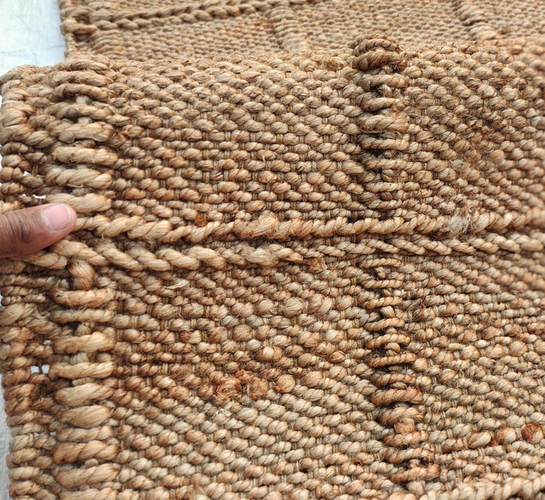 May include: A tan woven rug featuring a textured surface with a grid pattern created by thick, twisted fiber ropes. The material has a natural, earthy brown tone with a dense, knotted construction that forms a repeating square design throughout.