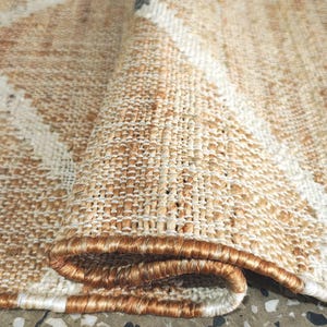 May include: A woven area rug featuring a natural tan fiber texture with a repeating white diamond pattern and small black accents at the intersections. The rug is folded over to reveal the thick, textured weave and a finished bound edge.