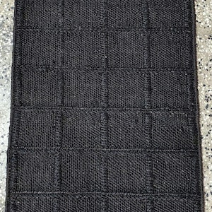 Puede incluir: Una alfombra rectangular negra tejida con un patr&oacute;n de cuadr&iacute;cula. La alfombra est&aacute; hecha de un material texturizado, posiblemente fibras naturales, y tiene un color oscuro y s&oacute;lido. El tejido crea un patr&oacute;n sutil de cuadrados y rect&aacute;ngulos.
