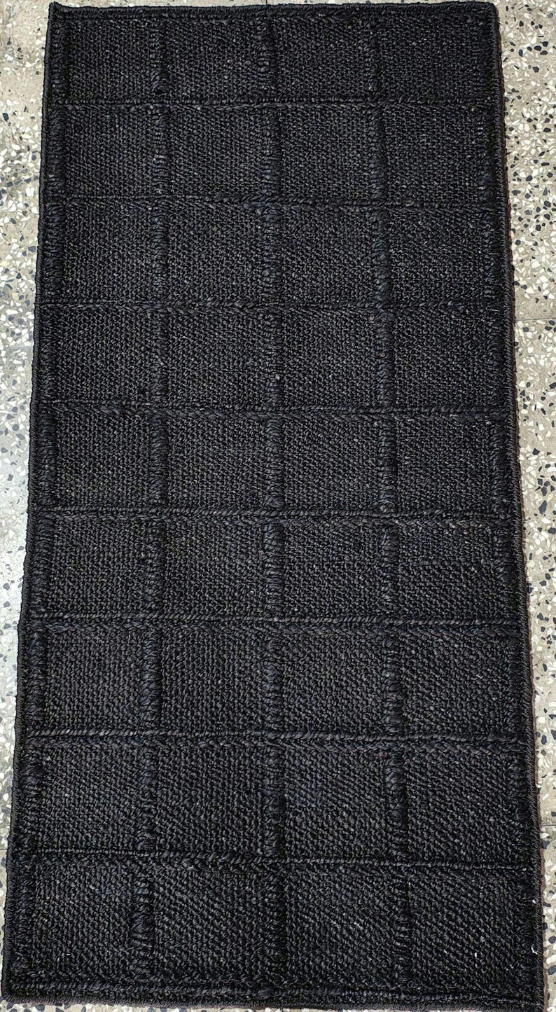 Puede incluir: Una alfombra rectangular negra tejida con un patr&oacute;n de cuadr&iacute;cula. La alfombra est&aacute; hecha de un material texturizado, posiblemente fibras naturales, y tiene un color oscuro y s&oacute;lido. El tejido crea un patr&oacute;n sutil de cuadrados y rect&aacute;ngulos.