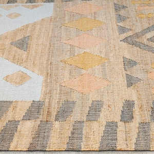 May include: A rectangular woven rug featuring a natural tan fiber base with geometric patterns in white, charcoal gray, mustard yellow, and soft peach. The design includes triangles, diamonds, and repeating block borders across the surface.
